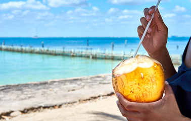Fresh coconut drink directly from the fruit / Hand of black woman holding coconut with drinking straw at caribbean coast