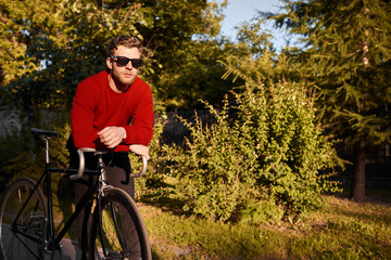 Naklejka premium Attractive bearded hipster in stylish clothes and sunglasses standing outdoors, leaning elbows on handlebar of his bike, having rest, enjoying beautiful nature around him. People and active lifestyle