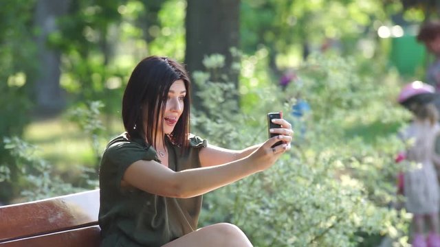 Girl doing selfie in the park The girl makes selfie. A girl is taking pictures of herself on the phone while sitting in a park on a bench