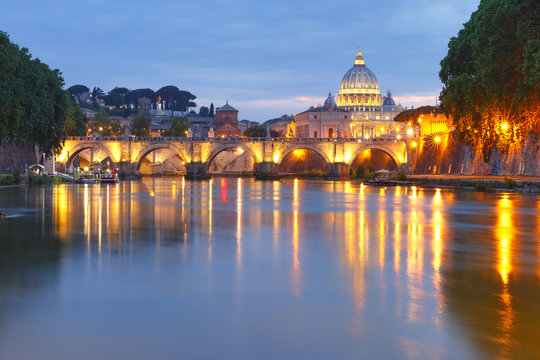 Saint Angel bridge and Saint Peter Cathedral with a mirror reflection in the Tiber River during evening blue hour in Rome, Italy.