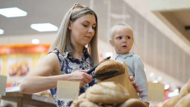 Happy attractive modern adult mommy with toddler kid pick up rolls with tongs from shelf in casual minimarket and put in craft package stand in aisle. Selection and buy fresh bread for breakfast