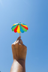 small colored paper cocktail umbrella on the beach, holding in his hand on blue sky background
