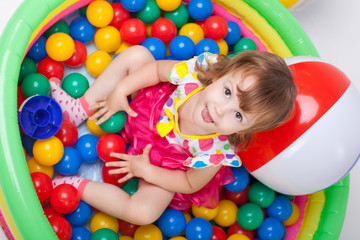 Fototapeta na wymiar маленькая девочка играет в бассейне с разноцветными шарами. Девочка показывает язык