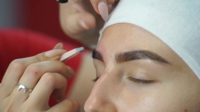 A young woman plucking eyebrows with tweezers. The master pulls the eyebrows from the client with tweezers. Procedures in the beauty salon