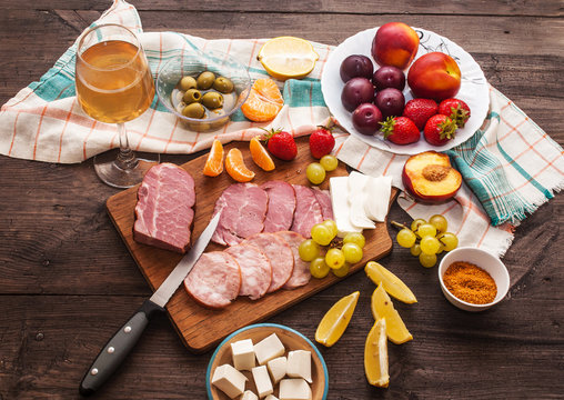 White wine and a set of meat delicacies, cheese and fruits on a dark, wooden background. Breakfast and cooking. View from above.