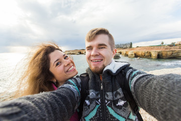 Young laughing couple hiking taking selfie with smart phone. Happy young man and woman taking self portrait with sea or ocean scenery on background. Winter time