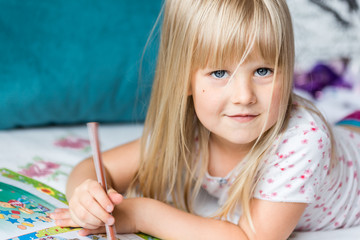 Portrait of cute little blonde girl lying on a bed and making homework in  a workbook with a pencil. End of vacation and back to school concept.