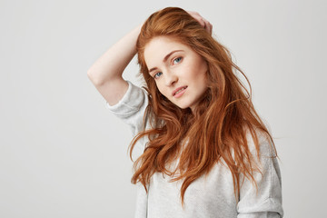 Portrait of young pretty ginger girl with freckles looking at camera smiling touching hair over white background.