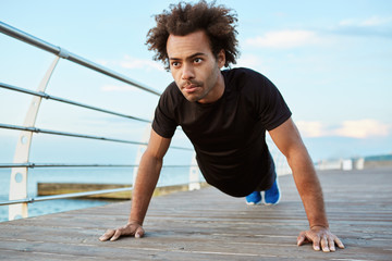 Fototapeta premium Motivated and concentrated Afro-American athlete with bushy hair wearing black running outfit standing in plank position on a wooden platform. Dark-skinned male runner preparing for morning workout
