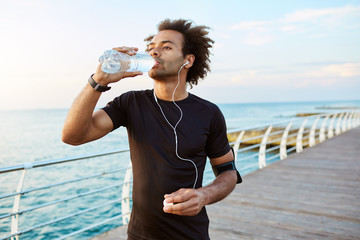 Stylish Afro-American male runner drinking water out of plastic bottle after cardio workout, wearing white earphones. Sportsman in black sportswear hydrating during outdoor training. - Powered by Adobe
