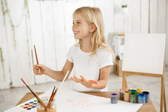 Smiling angel-like beautiful child with blonde hair wearing white t-shirt painting on her palm. Charming little girl drawing picture for her father, preparing birthday surprise for him. Children and