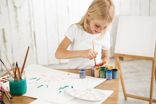 Focused and inspired little blonde girl deeping brush into paint, mixing it. Female freckled child in white t-shirt occupied with painting.