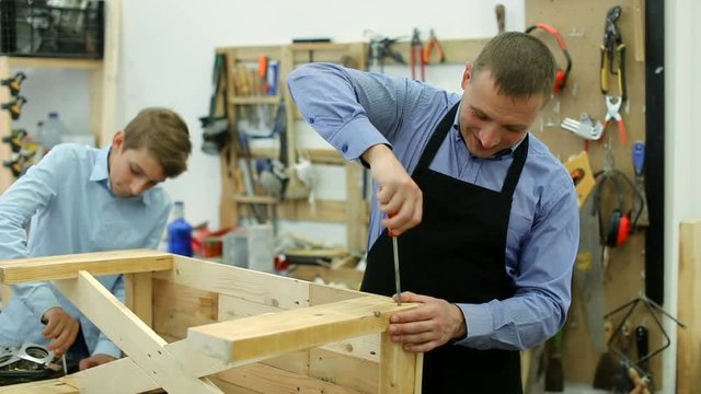 happy teacher and boy chiselling a wooden bench in workshop
