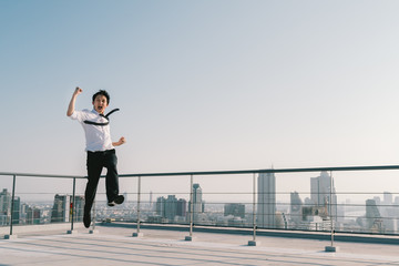 Fototapeta na wymiar Young handsome Asian businessman jumping high, celebrate success winning pose on building rooftop. Work, job, or successful business concept. Cityscape background with copy space on sunny blue sky