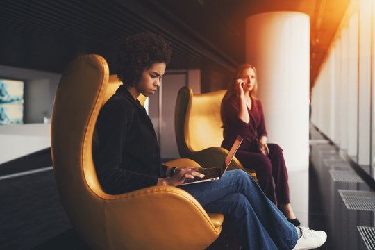 Young thoughtful afro american lady on yellow armchair is filling up table using laptop, her caucasian female colleague in defocused background having phone conversation, both are sitting near window