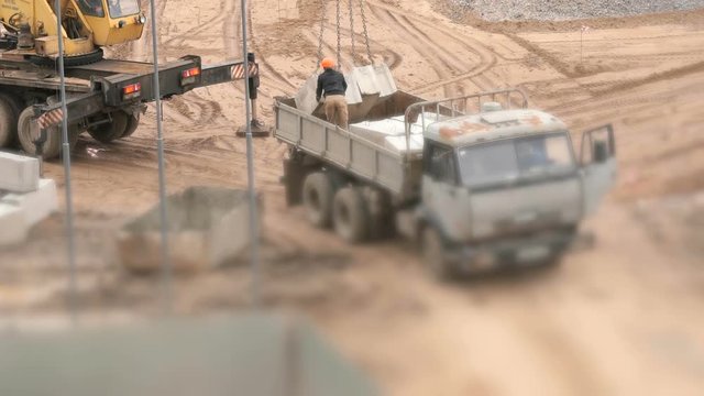 Builder dressed in orange helmet provides uploading of foundation blocks on the dump truck. Mobile construction crane on wheels