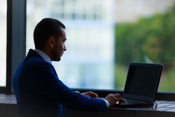 Portrait of young business man near big office window typing on laptop