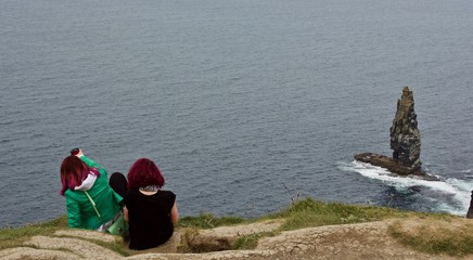 Dangerous Perch on Cliffs - two red-headed young women perch dangerously on the edge of the Cliffs of Moher, Ireland, during a gray spring day in 2017