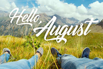 Hello august text with couple's legs together on view of the green valley and high mountains. Nature calendar background. Travel, vacation, holiday, unity with nature, adventure concept. - obrazy, fototapety, plakaty