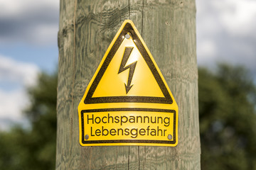 German high voltage sign mounted on wooden pole with green trees in background