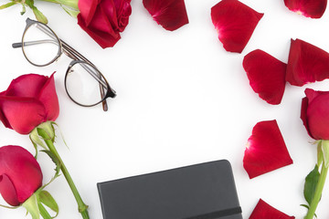 Black notebook and glasses decorated with red roses and petals on white background with copy space