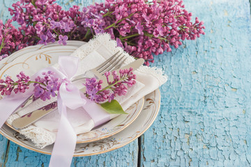 Tableware and silverware with bunches of violet lilac on the wooden background