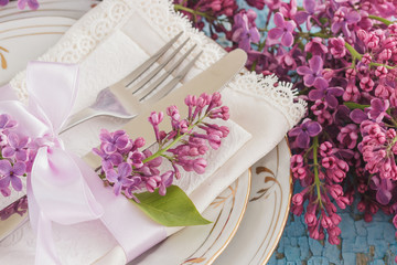Tableware and silverware with bunches of violet lilac on the wooden background