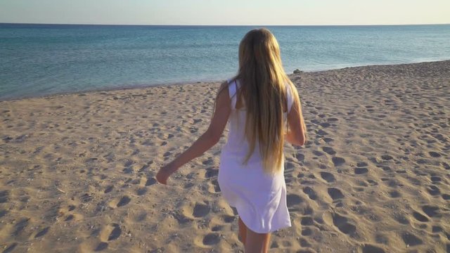 Young happy girl with long blonde hair running across sandy beach towards the sea. Barefoot teen girl in short white dress having fun during summer holidays vacation. Gimbal shot.