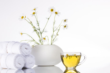 Bouquet of chamomiles flowers in a white vase and a transparent cup with tea on a white background. Towel, spa.