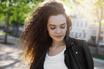 Beauty, fashion and style concept. Close up portrait of charming young brunette woman with long curls posing in urban setting, looking down with subtle smile, having a walk alone to make up her mind