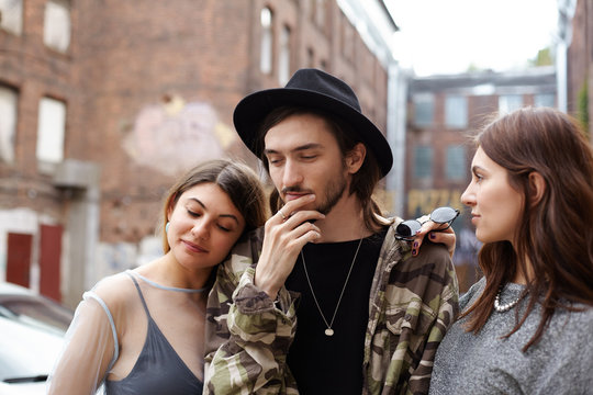 Stylish bearded guy having thoughtful look, holding hand in chin, facing dilemma as he has to choose between two gorgeous beautiful females who standing close to him, trying to attract his attention