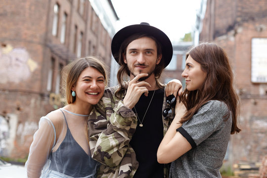 Attractive young European man wearing trendy hat and camouflage jacket relaxing in urban setting with his two girlfriends, enjoying good company. Three cheerful teenage friends having fun outdoor