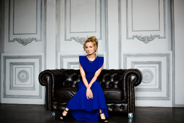 Красивая девушка с прической сидит на кожаном диване в синем вечернем платье в интерьерной студии - obrazy, fototapety, plakaty