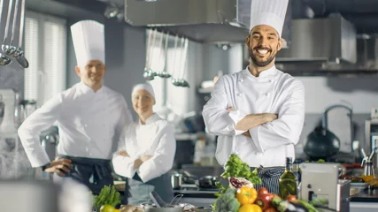 Poster Beroemde chef-kok van een groot restaurant kruist armen en glimlacht in een moderne keuken. Zijn staf in lachend op de achtergrond. © Gorodenkoff