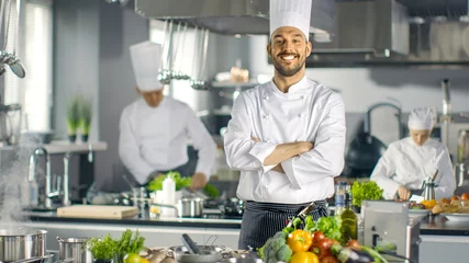 Fototapete Kochen Berühmter Koch eines großen Restaurants kreuzt Arme und lächelt in einer modernen Küche. Seine Mitarbeiter bei der Arbeit im Hintergrund.