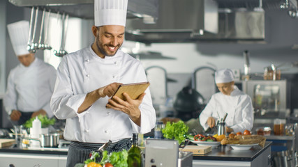 Famous Chef Uses Tablet Computer for Recipes While Working in a Modern Kitchen. His Help Work in...