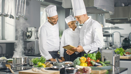 In het moderne keukenteam van koks gebruiken tabletcomputer voor recepten, ze glimlachen en hebben discussie. Keuken staat vol met voedselingrediënten, groenten, vlees, kokende soep.