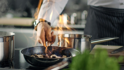 Le chef professionnel cuisine le style flambé. Il prépare un plat dans une casserole avec des flammes nues. Il travaille dans une cuisine moderne avec différents ingrédients qui traînent.