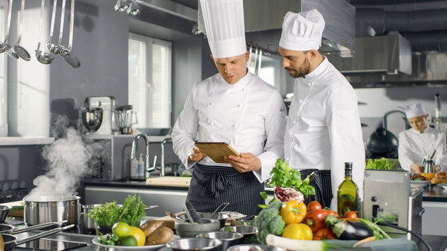 Two Famous Chefs Discuss Their Video Blog while Using Tablet Computer. They Work on a Big Restaurant Stainless Steel Professional Kitchen.