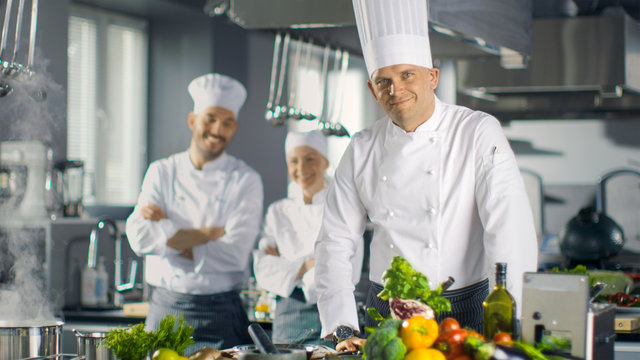 Famous Chef of a Big Restaurant Prepares Dishes and Smiles On Camera. In the Background Two Apprentices and Modern Kitchen.