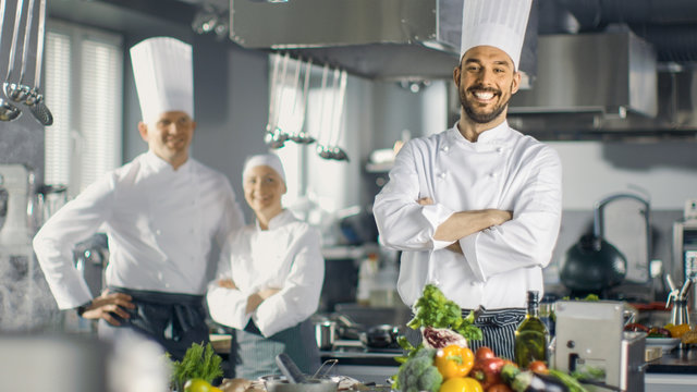 Famous Chef of a Big Restaurant Crosses Arms and Smiles in a Modern Kitchen. His Staff in Smiling in the Background.