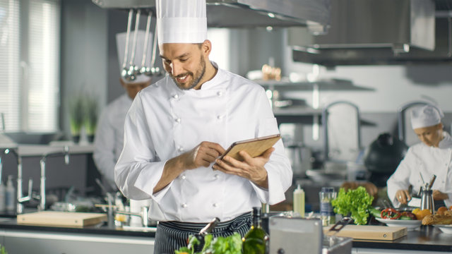 Famous Chef Uses Tablet Computer for Recipes While Working in a Modern Kitchen. His Help Work in the Background.