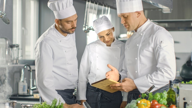 In the Modern Kitchen Team of Cooks Use Tablet Computer For Recipes, They Smile and Have Discussion. Kitchen is Full of Food Ingredients, Vegetables, Meat, Boiling Soup.