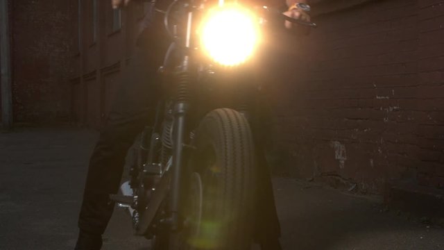 Black custom made cafe racer bike. Handsome man sitting on a motorbike, and starts it. The light from the headlights shining into the camera. Motorcycle leaves the frame.
