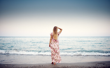 Beautiful portrait of stylish female in floral dress looking at blue water and ocean horizon at the dawn.