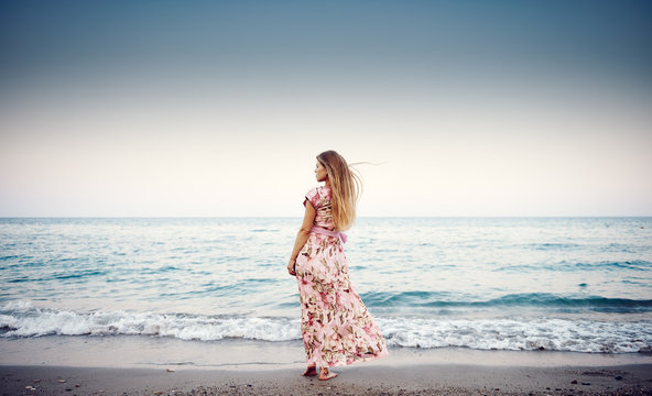 Young female in long dress standing on tropical coast enjoying ocean wind and sunset.