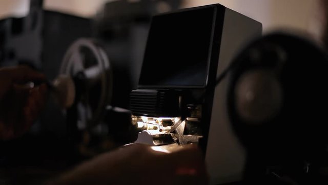 An editor moving the handles of a vintage film editing machine (a moviola, a device that allows viewing a reel on a tiny screen at a reduced speed), at a constant speed, in a dark room.
