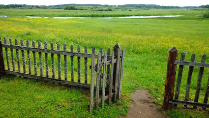 An open gate in a wooden fence and a green meadow beyond it, the path in the frame . Cloudy summer or late spring outside the city