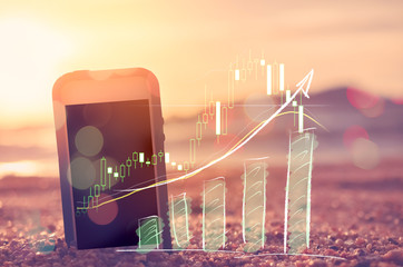 Business economic technology and freedom trader working concept. Smart phone on sand sunset beach double exposure graph money stock trading up trend arrow bar bokeh background.