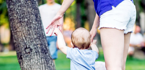 A little baby son walking supported by his mother to meet a father in summer park